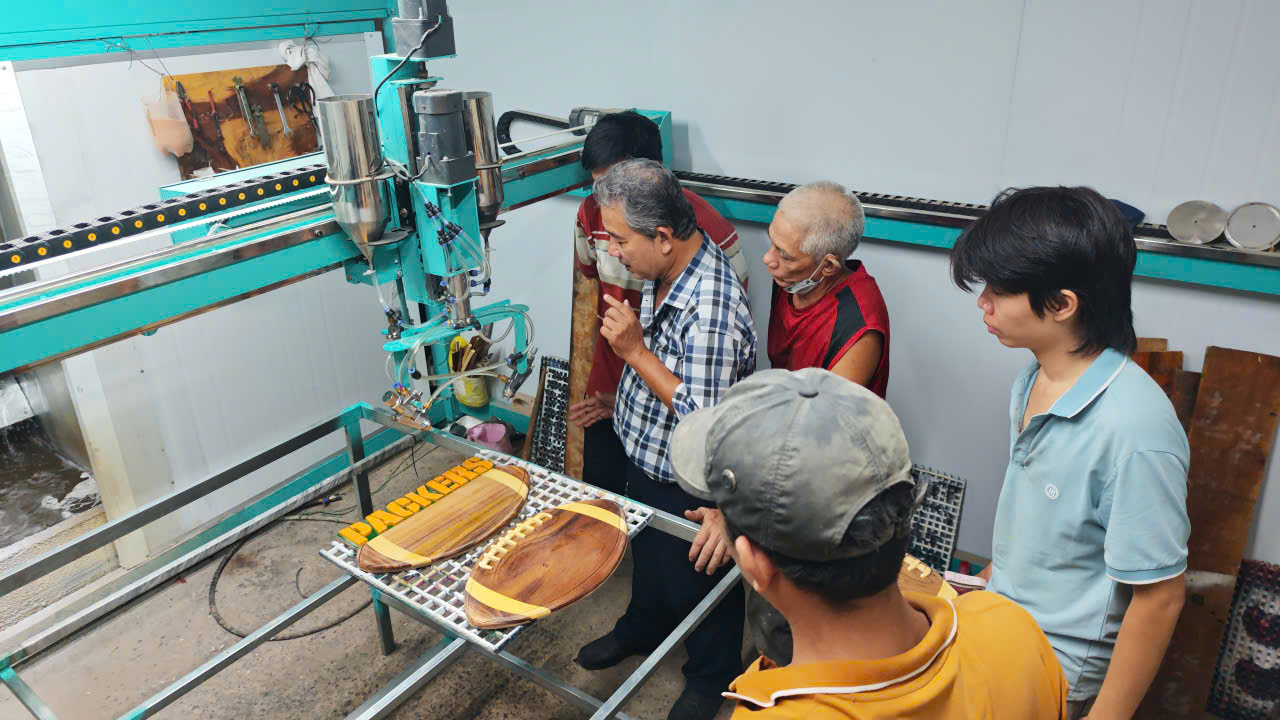 Máy phun sơn tự động khung xanh với hai bình chứa sơn đang phun các mẫu đồ gỗ lưu niệm tại xưởng