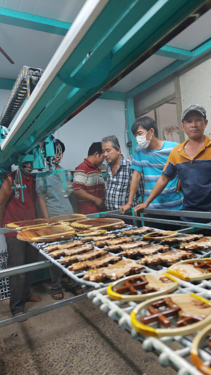 Nhóm kỹ thuật và chủ xưởng cùng quan sát máy phun sơn tự động vận hành trên giá đồ gỗ lưu niệm